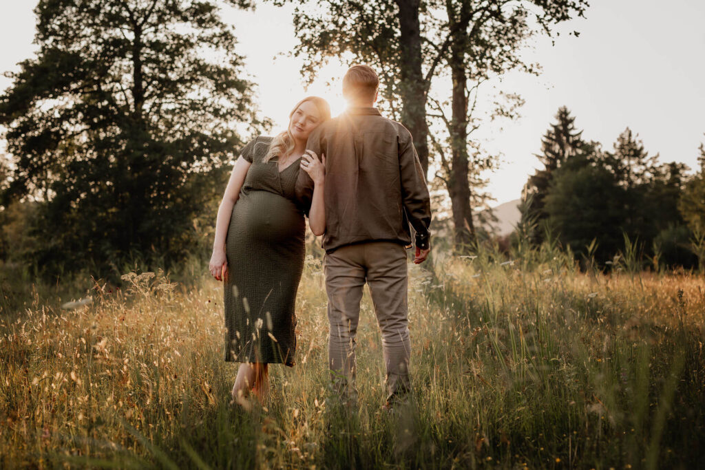 Eine schwangere Frau lehnt sich bei einem Babybauch Fotoshooting an die Schulter ihres Mannes an. Das Bild wurde bei Sonnenuntergang am Mondsee in einer wilden Wiese gemacht.