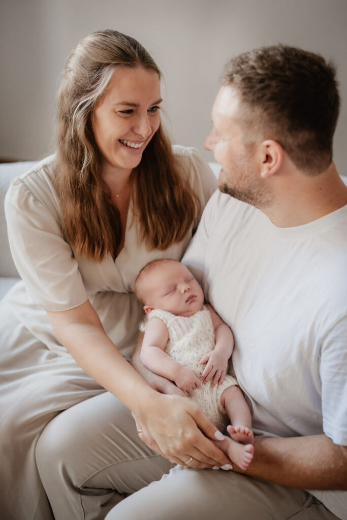 Eltern bei einem Newbornshooting lachen sich entspannt an. Auf ihren Knien liegt ein Neugeborenes und schläft.