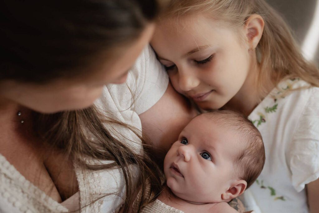 Eine kleine Familie hat sich bei einem Newborn-Fotoshooting um ein neugeborenes Baby versammelt und schaut es liebevoll an.